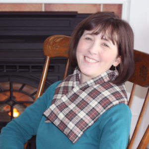 Extra long microwaveable neck wrap (in black and cream colored plaid flannel) helping a woman relax stiff muscles near wood stove