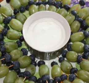 Green grapes and blueberries on toothpicks arranged around a plate with yogurt dip in the center